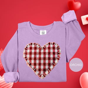 May include: A lilac sweatshirt with a heart-shaped patch in a red and white gingham pattern and a ruffled edge. The word "ORCHID" is in a circle on the right. The background is red with heart decorations. The sweatshirt is a Comfort Colors brand.
