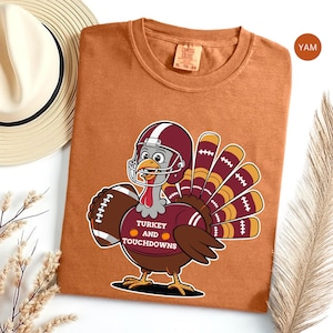 Könnte beinhalten: Ein rostorangefarbenes T-Shirt mit einem Cartoon-Truthahn, der einen kastanienbraunen Football-Helm und ein Trikot trägt. Der Truthahn hält einen Football und der Text auf dem Trikot lautet TURKEY AND TOUCHDOWNS mit zwei kleinen orangefarbenen Kürbissen.
