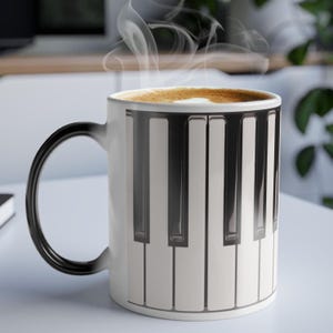 以下が含まれることがあります： 黒い持ち手が付いた白いセラミックコーヒーマグ。ピアノのキーボードのデザインが施されています。マグカップには温かい飲み物が入っており、上部から湯気が立ち上っています。マグカップは白い表面に置かれています。