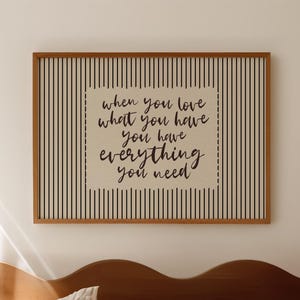 May include: A rectangular framed wall art piece featuring a vertical black pinstripe pattern on a cream background. Centered is a cream-coloured rectangular box with the black text: when you love what you have you have everything you need.