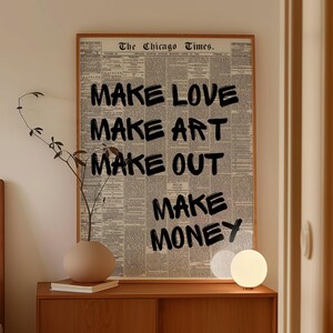 以下が含まれることがあります： ヴィンテージ新聞を背景に、黒で「MAKE LOVE MAKE ART MAKE OUT MAKE MONEY」と書かれた額入りのプリント。木製のキャビネットの上に飾られており、ベージュの花瓶と小さな丸いランプが置かれています。