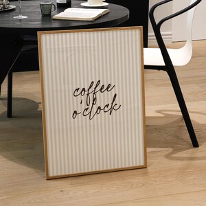 May include: A framed vertical sign with the text "coffee o'clock" in cursive script. The sign has a light brown wooden frame and a striped background of beige and white. A black round table with a wine bottle, coffee cup, and book is in the background.
