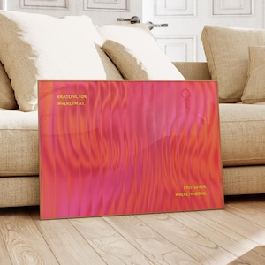 May include: A rectangular art print with a vibrant abstract design in shades of pink and orange. The print features the phrases "Grateful for where I'm at" and "Excited for where I'm going" in gold lettering. The artwork is displayed on a wooden floor.