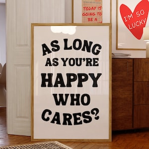 May include: A black and white poster with the text "As long as you're happy who cares?" in a bold, playful font. The poster is framed in a light wood frame and is hanging on a white wall.