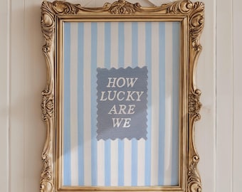 May include: A framed artwork with a gold ornate frame is hanging on a white wooden wall. The artwork features a blue and white striped background with a blue rectangle in the center that reads "HOW LUCKY ARE WE". A white bow is attached to the top.