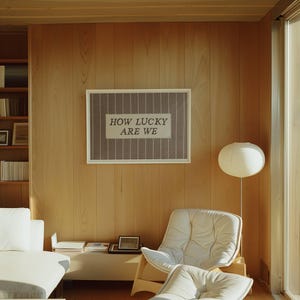 Puede incluir: Una obra de arte enmarcada con el texto "HOW LUCKY ARE WE" cuelga de una pared de madera clara. La obra presenta un fondo a rayas grises y blancas. Tambi&eacute;n se ven una silla blanca y una l&aacute;mpara de pie con pantalla redonda.