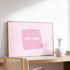 May include: A framed pink poster with a pink sticky note that says "I got this!" in white text. The poster is hanging on a white wall above a wooden desk with a chair.