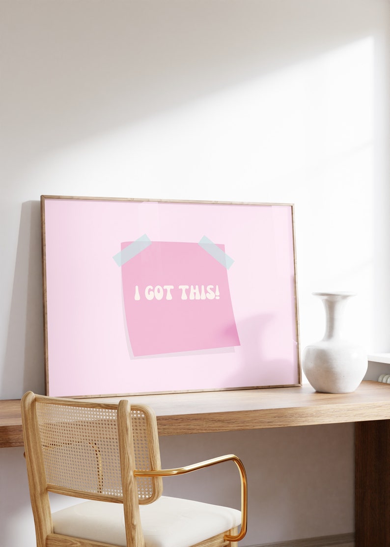 May include: A framed pink poster with a pink sticky note that says "I got this!" in white text. The poster is hanging on a white wall above a wooden desk with a chair.