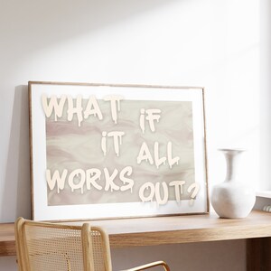 May include: A framed print with the text "WHAT IF IT ALL WORKS OUT?" in white paint on a light pink and white marbled background. The print is hanging on a white wall above a wooden desk with a woven chair and a white vase.