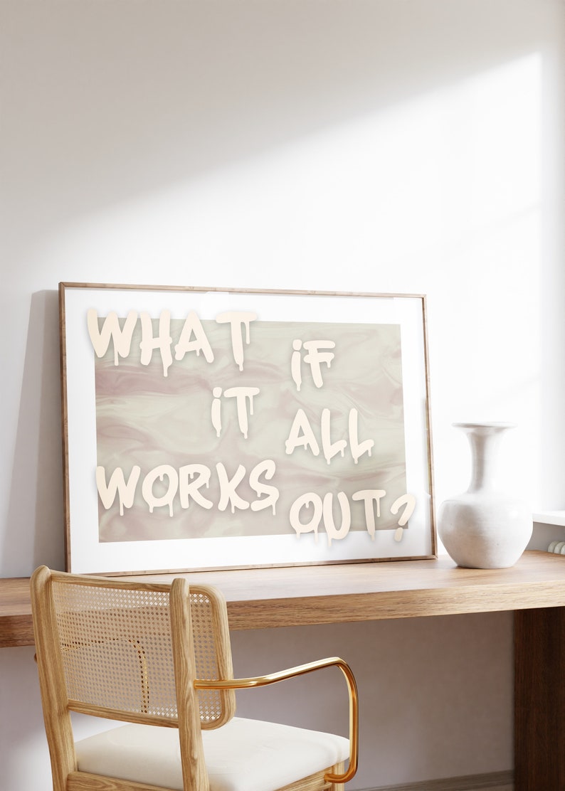 May include: A framed print with the text "WHAT IF IT ALL WORKS OUT?" in white paint on a light pink and white marbled background. The print is hanging on a white wall above a wooden desk with a woven chair and a white vase.