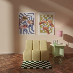 May include: Two framed prints with illustrations of hands holding glasses of cocktails. The prints are hanging on a light peach wall above a yellow modular chair and a light green side table with a pink lamp.