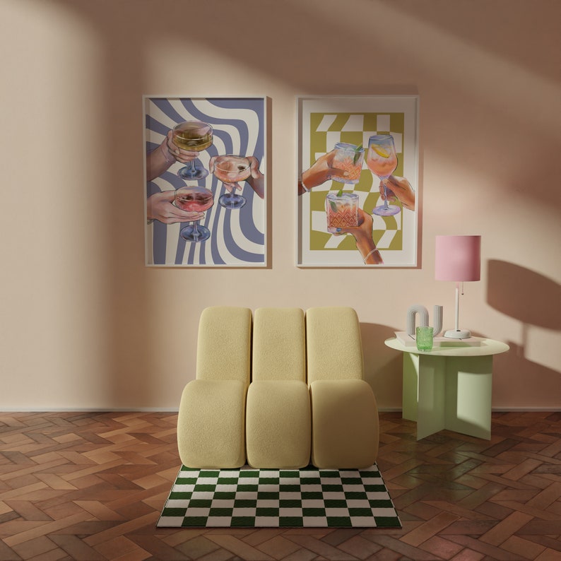 May include: Two framed prints with illustrations of hands holding glasses of cocktails. The prints are hanging on a light peach wall above a yellow modular chair and a light green side table with a pink lamp.