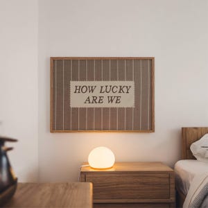 Puede incluir: Un cuadro enmarcado con el texto "HOW LUCKY ARE WE" cuelga sobre una mesita de noche de madera. Una l&aacute;mpara esf&eacute;rica emite un brillo c&aacute;lido. La habitaci&oacute;n tiene una paleta de colores neutros, con una cama visible al fondo.