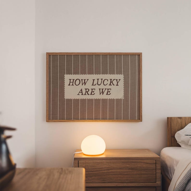 Puede incluir: Un cuadro enmarcado con el texto "HOW LUCKY ARE WE" cuelga sobre una mesita de noche de madera. Una l&aacute;mpara esf&eacute;rica emite un brillo c&aacute;lido. La habitaci&oacute;n tiene una paleta de colores neutros, con una cama visible al fondo.