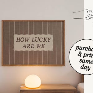 Puede incluir: Una obra de arte enmarcada con el texto "HOW LUCKY ARE WE" sobre un fondo a rayas. La obra est&aacute; en un marco de madera y cuelga de una pared blanca. Una l&aacute;mpara redonda est&aacute; sobre una mesita de noche de madera debajo. Texto a la derecha: "Made with Love - Arta".