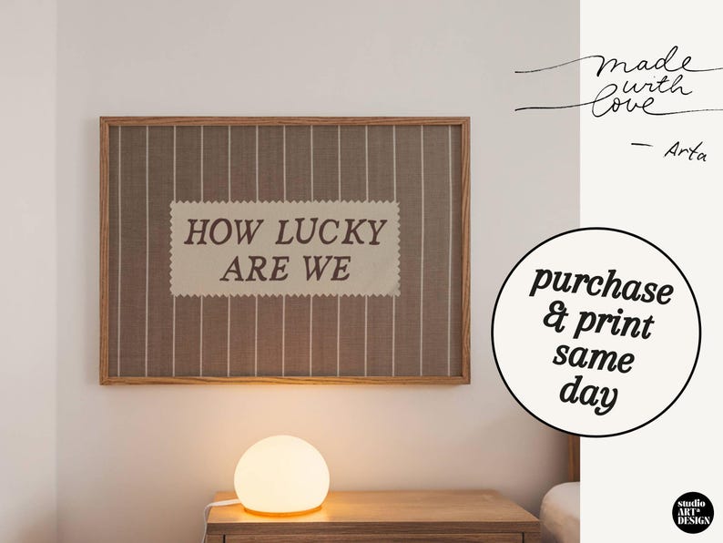 Puede incluir: Una obra de arte enmarcada con el texto "HOW LUCKY ARE WE" sobre un fondo a rayas. La obra est&aacute; en un marco de madera y cuelga de una pared blanca. Una l&aacute;mpara redonda est&aacute; sobre una mesita de noche de madera debajo. Texto a la derecha: "Made with Love - Arta".