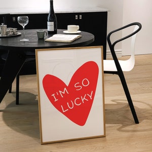 May include: A framed print with a red heart shape and the text "I'm so lucky" in white. The print is on a white background and has a wooden frame.