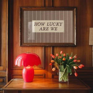 Puede incluir: Una l&aacute;mpara de mesa roja en forma de hongo con borde festoneado sobre una mesa de madera. Encima, una obra de arte enmarcada muestra el texto &laquo;HOW LUCKY ARE WE&raquo;. Un jarr&oacute;n de tulipanes a&ntilde;ade color.