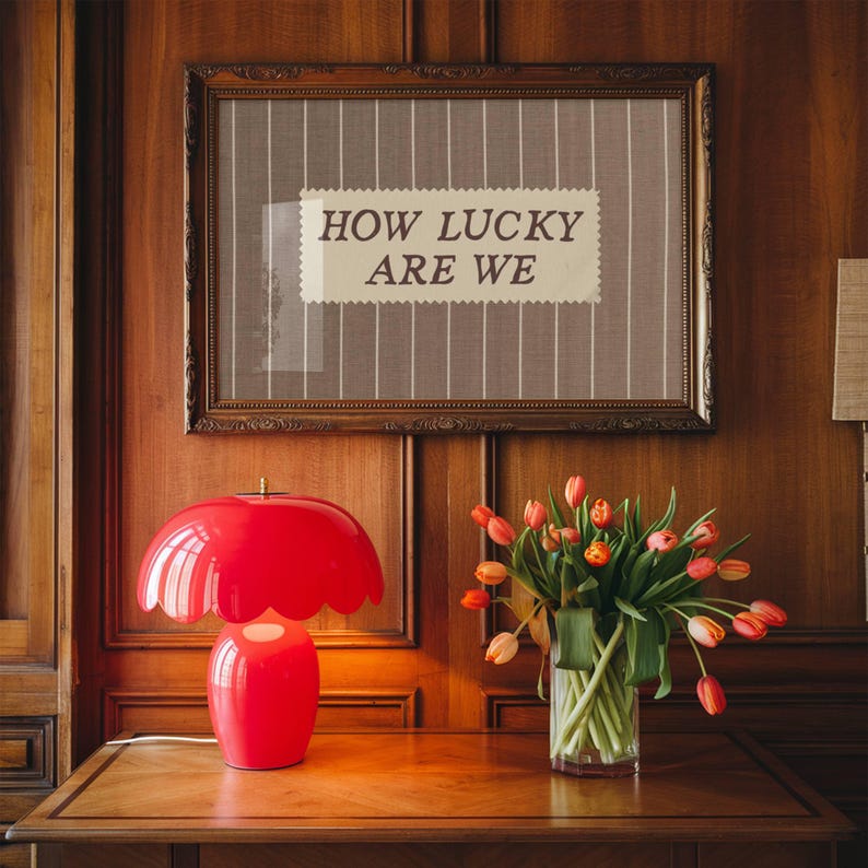 Puede incluir: Una l&aacute;mpara de mesa roja en forma de hongo con borde festoneado sobre una mesa de madera. Encima, una obra de arte enmarcada muestra el texto &laquo;HOW LUCKY ARE WE&raquo;. Un jarr&oacute;n de tulipanes a&ntilde;ade color.