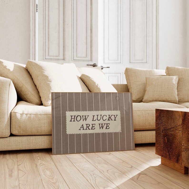 Puede incluir: Un letrero rectangular con el texto "HOW LUCKY ARE WE" en blanco, con un estilo vintage. El letrero tiene un fondo a rayas grises y blancas y est&aacute; colocado frente a un sof&aacute; beige con cojines a juego.