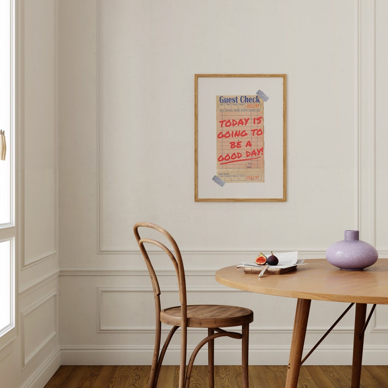 May include: A framed print with a vintage guest check that says "TODAY IS GOING TO BE A GOOD DAY!" hanging on a white wall above a wooden table and chair.