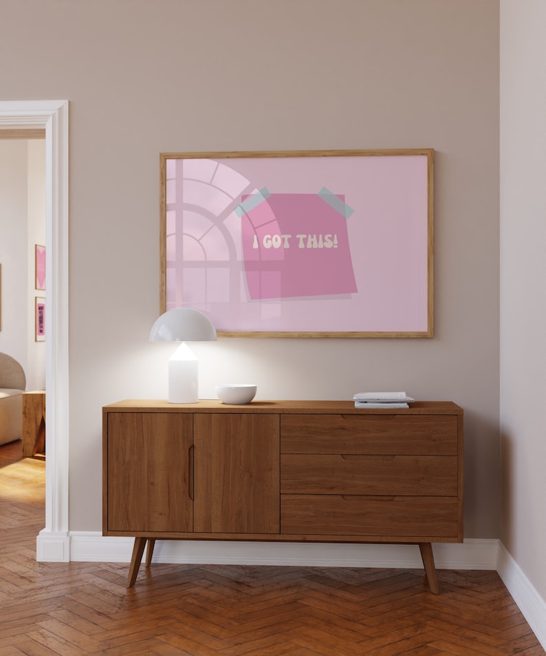 May include: A light wood sideboard with two doors and three drawers. A white lamp and a white bowl are on top of the sideboard. A framed print with a pink background and a pink sticky note with the text "I GOT THIS!" is hanging on the wall above the sideboard.