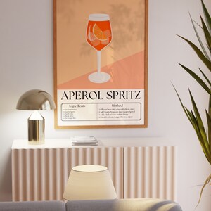 May include: A printable poster with an illustration of an Aperol Spritz cocktail. The poster has a peach background with a brown wood frame. The text on the poster reads "Aperol Spritz" and includes a list of ingredients and instructions for making the cocktail.