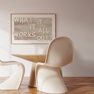 May include: A framed print with the text "WHAT IF IT ALL WORKS OUT?" in white paint on a beige background. Two white chairs and a wooden table are in the foreground.