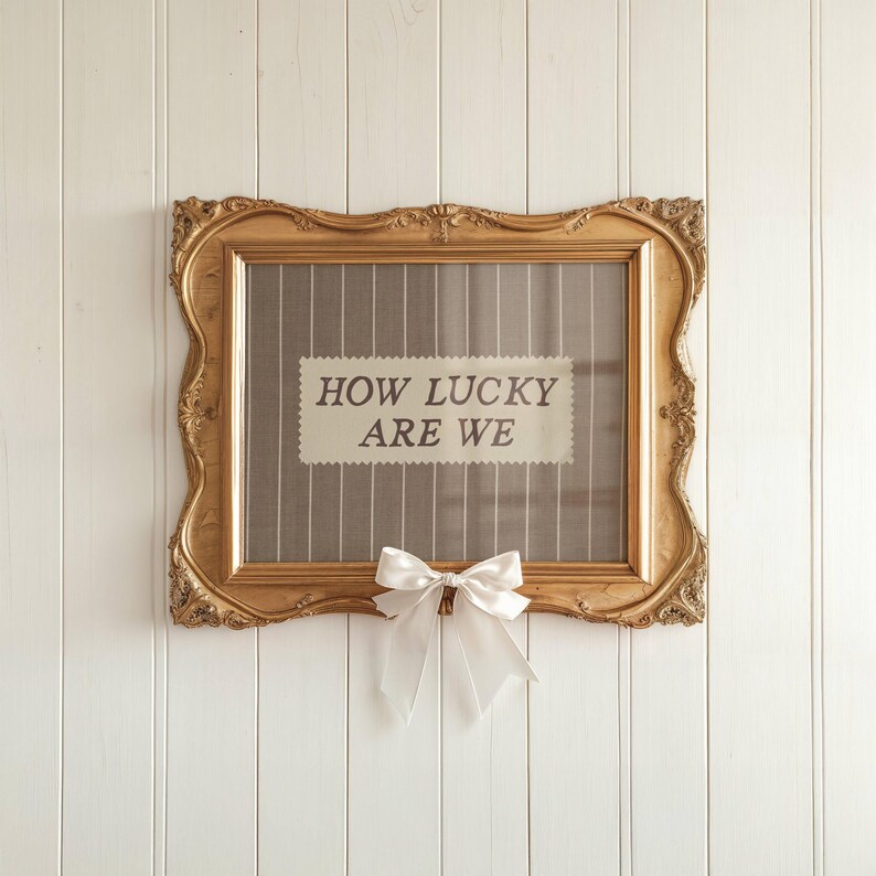 Puede incluir: Un marco dorado con la frase "HOW LUCKY ARE WE" en una etiqueta rectangular. El marco tiene detalles ornamentales y un lazo blanco en la parte inferior. El fondo es un panel de madera vertical blanco.