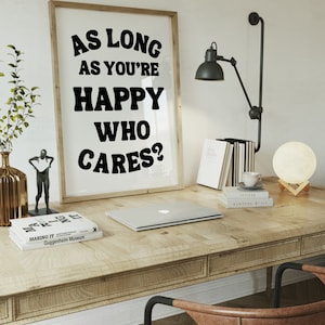 May include: A framed print with the text "As long as you're happy who cares?" in black bold letters on a white background. The print is hanging on a white wall above a wooden desk with a laptop, books, and a lamp.