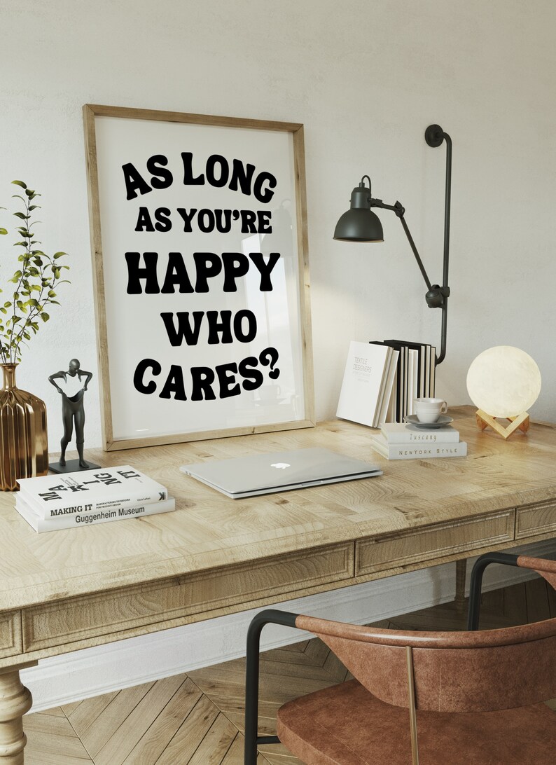 May include: A framed print with the text "As long as you're happy who cares?" in black bold letters on a white background. The print is hanging on a white wall above a wooden desk with a laptop, books, and a lamp.
