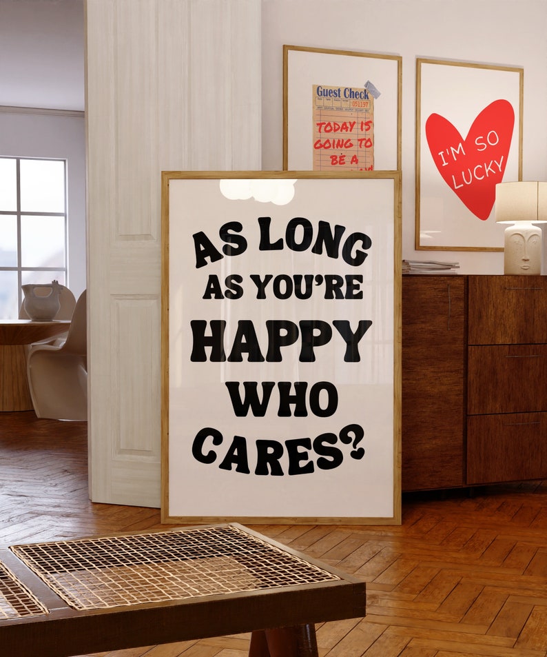 May include: A black and white poster with the text "As long as you're happy who cares?" in a bold, playful font. The poster is framed in a light wood frame and is hanging on a white wall.