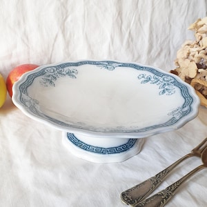 Puede incluir: Un soporte para tartas de cerámica blanca con un diseño floral azul y blanco. El soporte tiene un borde festoneado y una base de pedestal.