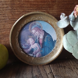 May include: A gold-framed picture with a religious scene depicting a mother and child. The mother is wearing a blue robe and the child is wearing a white robe. The picture is set against a wooden background.