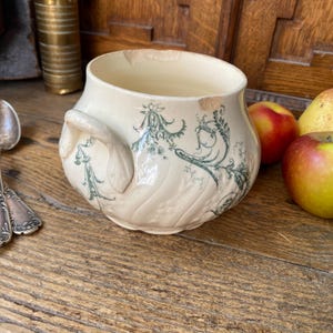 Puede incluir: Cuenco de cerámica antigua de color crema con asa y diseño floral verde. El cuenco tiene un borde ligeramente astillado y está sobre una superficie de madera. Hay manzanas y una pera junto al cuenco.