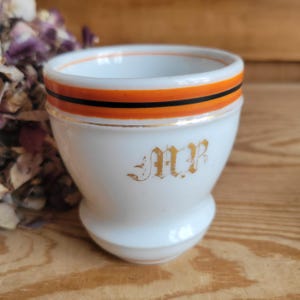 May include: A white porcelain egg cup featuring a wide orange band with a thin black stripe and a gold rim. The front of the cup displays the gold-colored monogram letters MP in a decorative gothic-style script font, centered on the smooth surface.