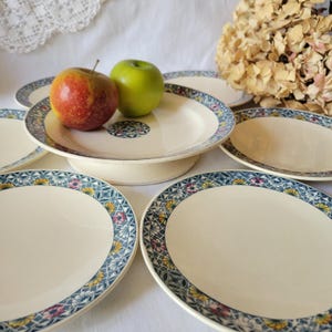 May include: Set of vintage ceramic plates with a blue and white floral pattern. The plates have a raised rim and a center circle with a floral design. The plates are arranged in a stack with two apples on top.