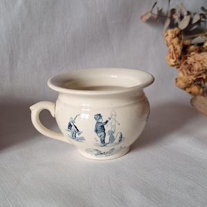 May include: Cream-colored ceramic chamber pot with a handle and a wide rim. The pot is decorated with blue illustrations of children playing. A vintage, antique item.