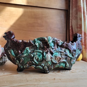 May include: An antique, oblong ceramic planter with a detailed relief design. The planter features a dark brown and green color scheme with ornate carvings of mythical creatures and foliage. It has a scalloped rim and is supported by small feet.