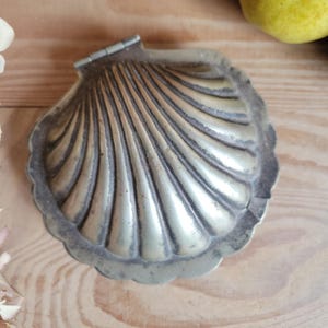 Puede incluir: Una caja de baratijas plateada con forma de concha con bisagras. La caja tiene un diseño detallado y acanalado y un borde festoneado. La caja es de metal y está abierta, revelando el interior.