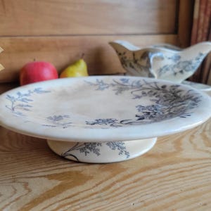 May include: A cream-colored ceramic pedestal plate featuring a dark blue floral pattern along the rim and base. The plate has a wide, flat surface and a sturdy, circular pedestal stand. The design includes intricate botanical illustrations.