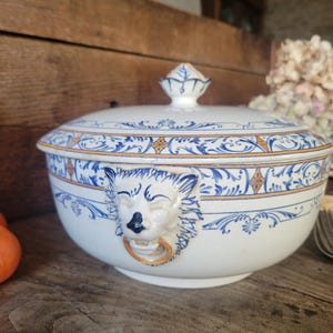 Könnte beinhalten: Weiße Keramik-Suppenterrine mit Deckel, verziert mit blauen und orangefarbenen Blumenmustern. Die Terrine hat einen Löwenkopfgriff mit goldenem Ring. Ein dekoratives Stück zum Servieren von Suppen und Eintöpfen.