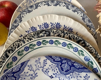 Op de afbeelding: Een stapel antieke witte keramische borden met blauwe bloemen- en krulwerkontwerpen. De borden hebben verschillende patronen en maten, sommige met geschulpte randen. Een rode appel en een gele citroen zijn zichtbaar.