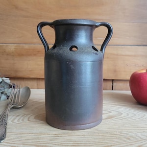 Può includere: Un vaso in ceramica marrone scuro con due manici e diversi piccoli fori vicino alla parte superiore. Il vaso ha una forma cilindrica e una base marrone leggermente più chiara. Lo sfondo include una superficie in legno, una forchetta, un cucchiaio e una mela rossa.