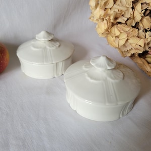 Op de afbeelding: Twee witte keramische doosjes met deksel en een decoratief bloemmotief. De doosjes zijn rond en hebben een verhoogd midden met een bloemmotief.