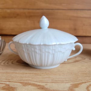 Puede incluir: Un cuenco blanco de porcelana con tapa y diseño de tejido de cesta texturizado. El cuenco tiene dos asas pequeñas y un adorno decorativo en la tapa. El cuenco está sobre una superficie de madera marrón claro.