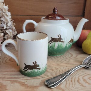 Könnte beinhalten: Ein weißes Keramik-Tassen- und Teekannen-Set mit einem braunen Hasen-Design. Die Tasse und die Teekanne haben einen verwitterten, Vintage-Look mit braunem Rand. Die Teekanne hat einen braunen Deckel und Ausguss. Das Set steht auf einer Holzoberfläche.