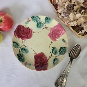 May include: A vintage ceramic plate with a floral design. The plate features four pink and red roses with green leaves on a cream background.