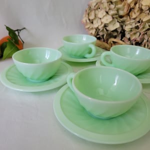 May include: A set of four light green teacups and saucers. The cups have a spiral design and small handles. The saucers are round with a matching pattern. The set is made of glass and is a vintage style.