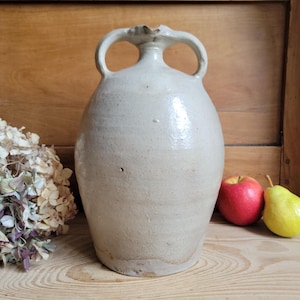 May include: A large, off-white stoneware jug with two arched handles. The jug has a rounded body and a slightly flared neck. A red apple and a yellow pear sit beside the jug. Dried flowers are on the left.